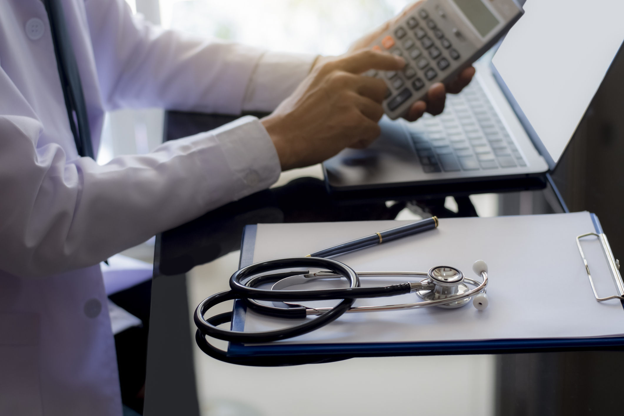 Male doctor or physician using calculator and work on laptop computer with medical stethoscope and clipboard on the desk at clinic or hospital. Medical healthcare costs ,fees and revenue concept that bicycle accidents lawyer assist obtain compensation for brain injuries